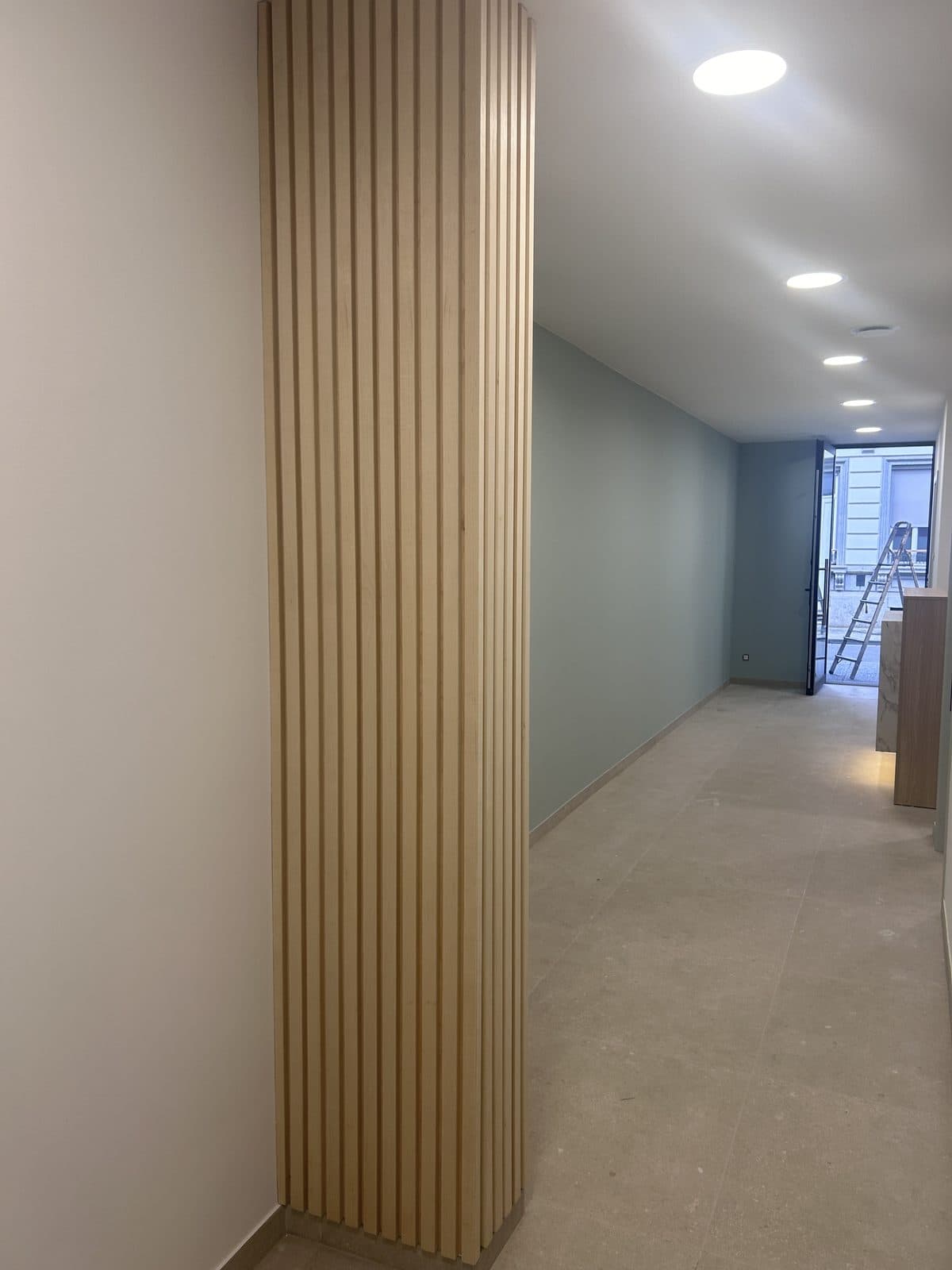 Couloir d’accueil : poteau habillé de lattes verticales en bois clair, mur vert sauge, sol en pierre, porte d’entrée vitrée