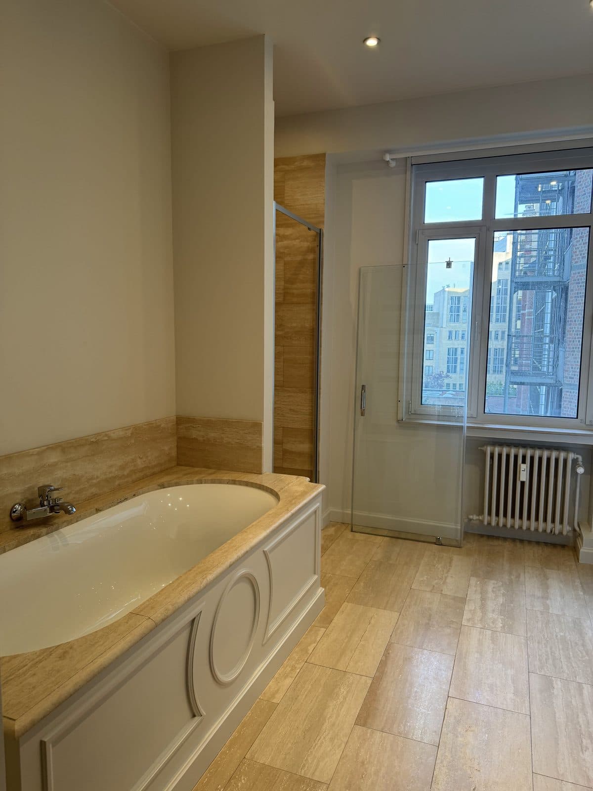 Salle de bain aspect travertin : baignoire à tablier mouluré, haute fenêtre sur la ville, radiateur en fonte, douche vitrée