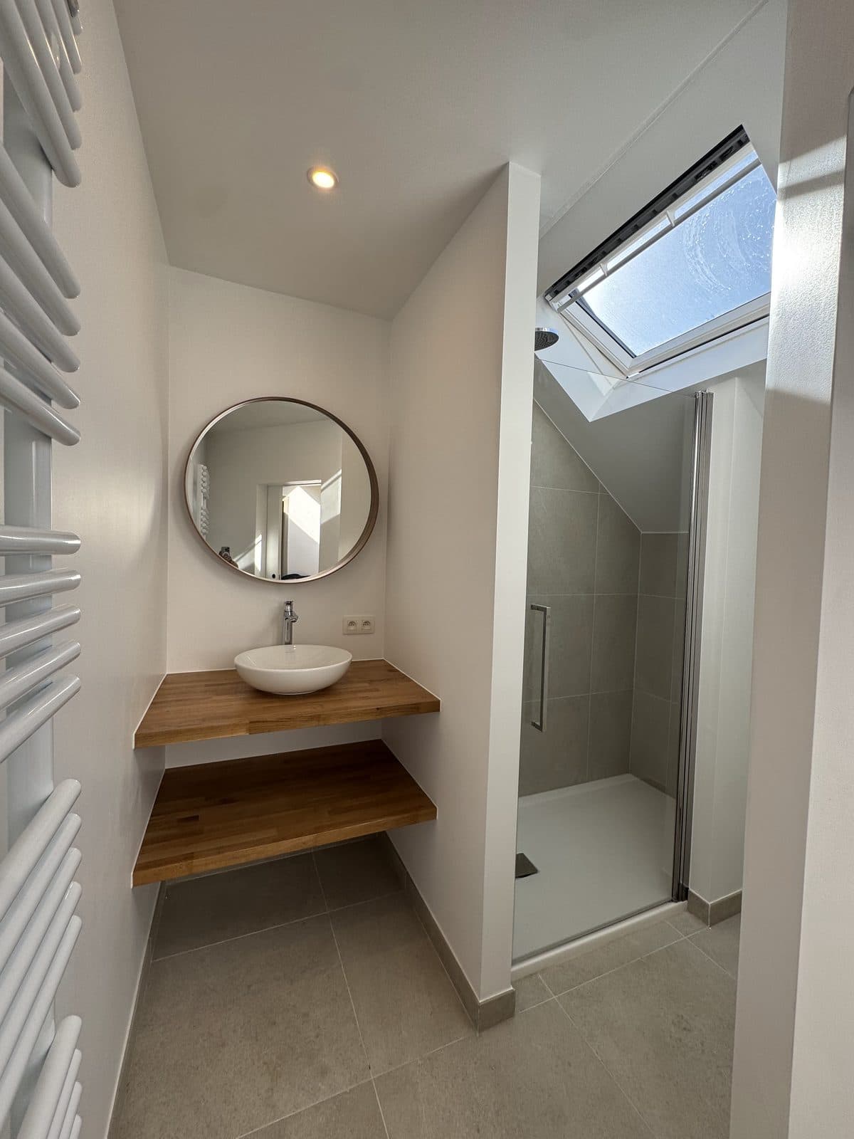 Petite salle de douche sous les toits : miroir rond, vasque à poser sur une tablette en chêne, douche sous une fenêtre de toit
