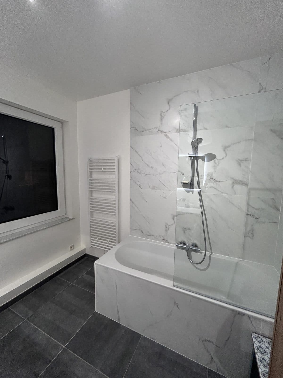 Baignoire à pare-douche vitré et colonne chromée, carrelage aspect marbre, sol foncé, sèche-serviettes blanc