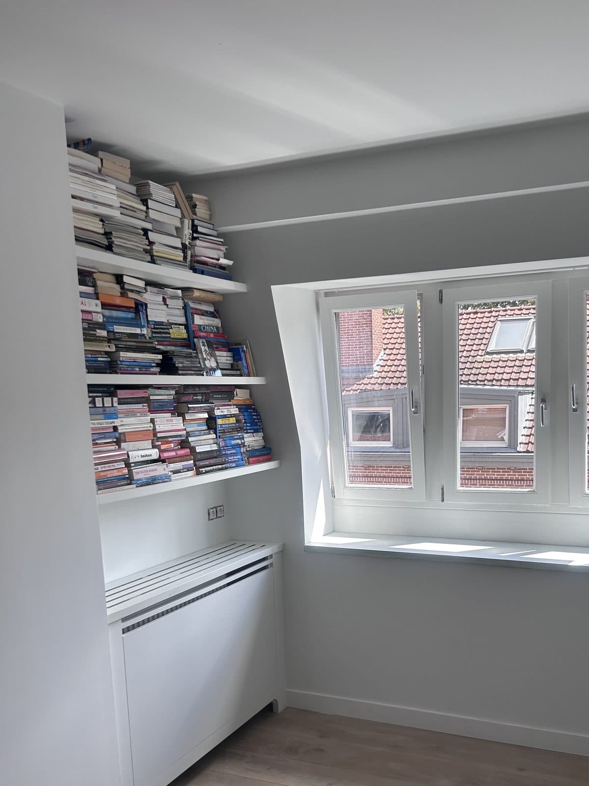 Cache-radiateur blanc sous des étagères blanches garnies de livres, lucarne sur des toits de tuiles rouges