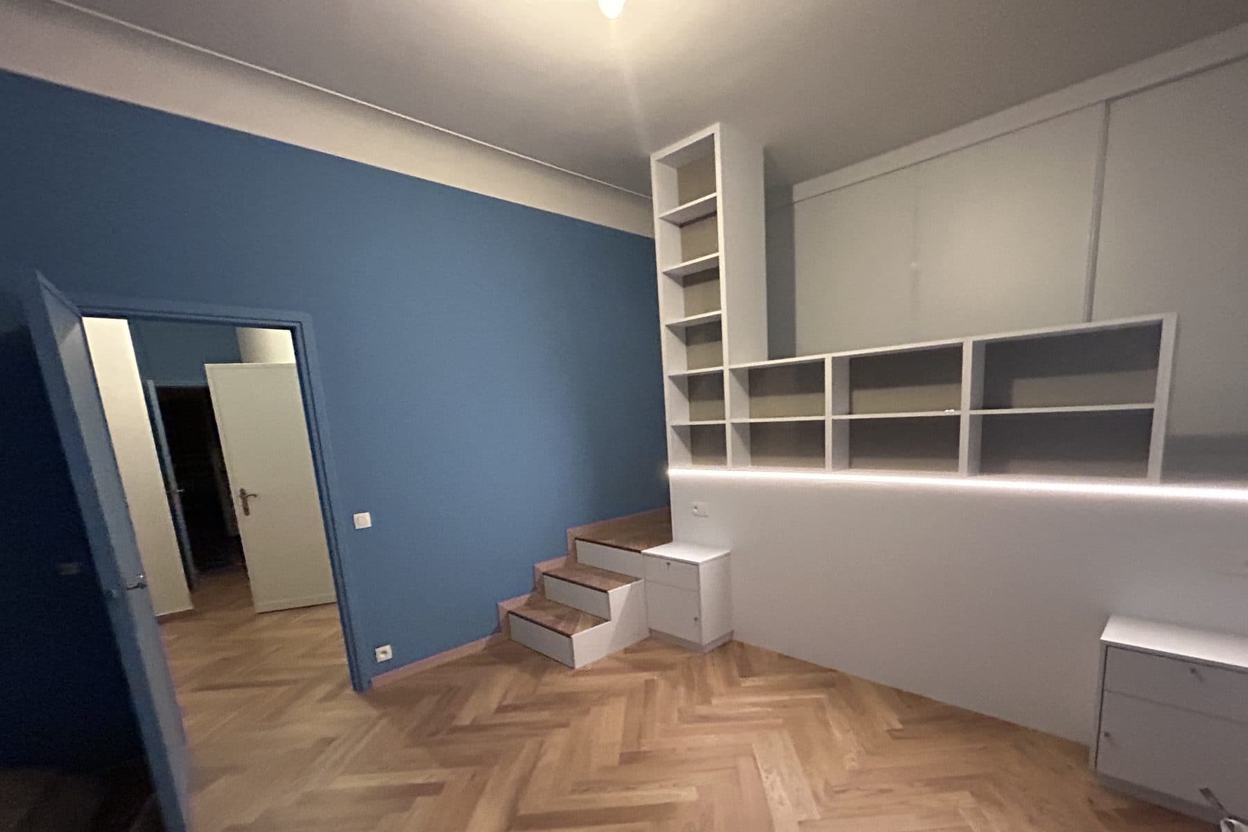 Chambre bleue vide : parquet à bâtons rompus, bibliothèque blanche intégrée à ligne lumineuse, marches à tiroirs menant à une estrade
