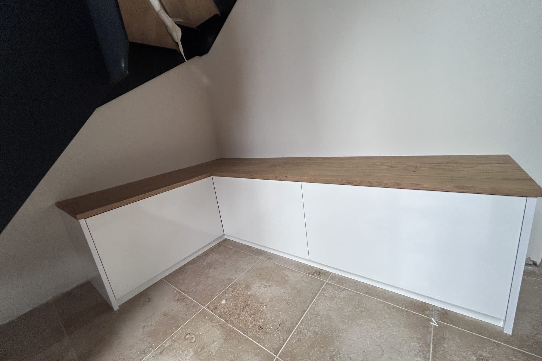 Banc bas en L sous un escalier noir, façades blanches laquées, dessus en chêne, carrelage en pierre beige