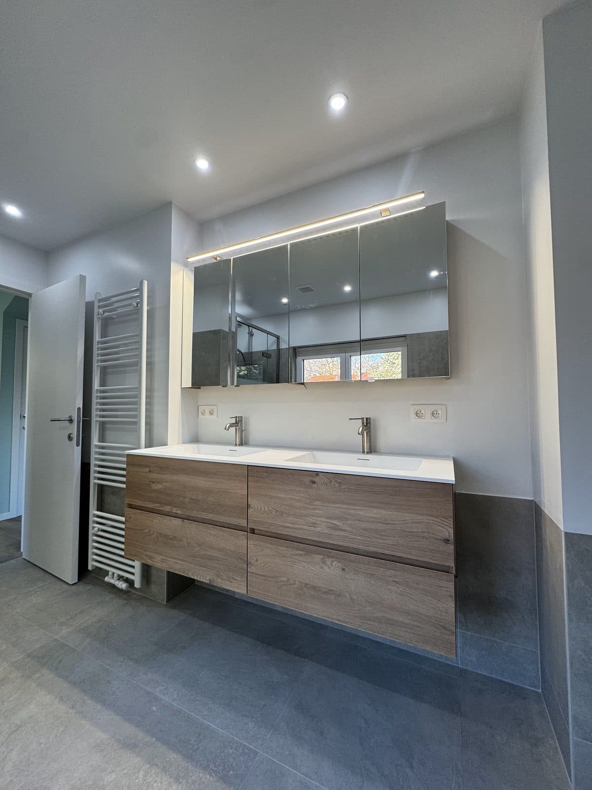 Salle de bain : carrelage gris, meuble double vasque suspendu en bois, armoire miroir éclairée, sèche-serviettes