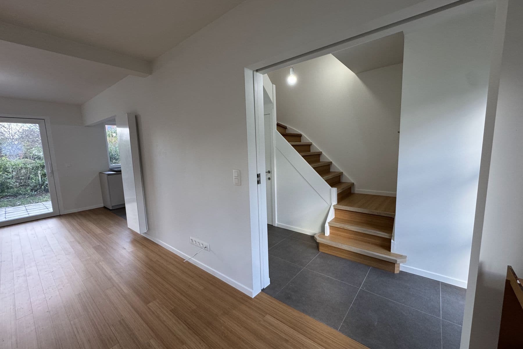 Pièce livrée : parquet ton bambou, carrelage gris foncé devant l’escalier, porte-fenêtre sur le jardin