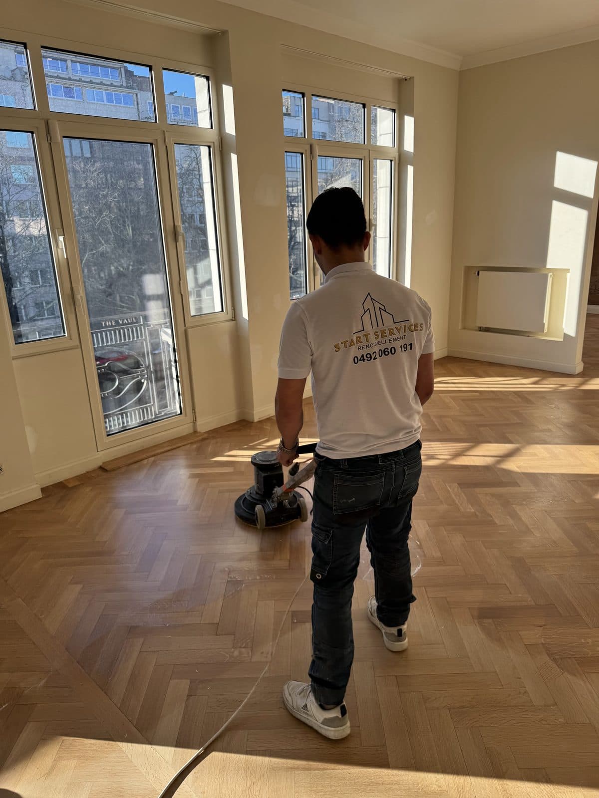 Un ouvrier, de dos, passe la monobrosse sur un parquet à bâtons rompus, hautes fenêtres