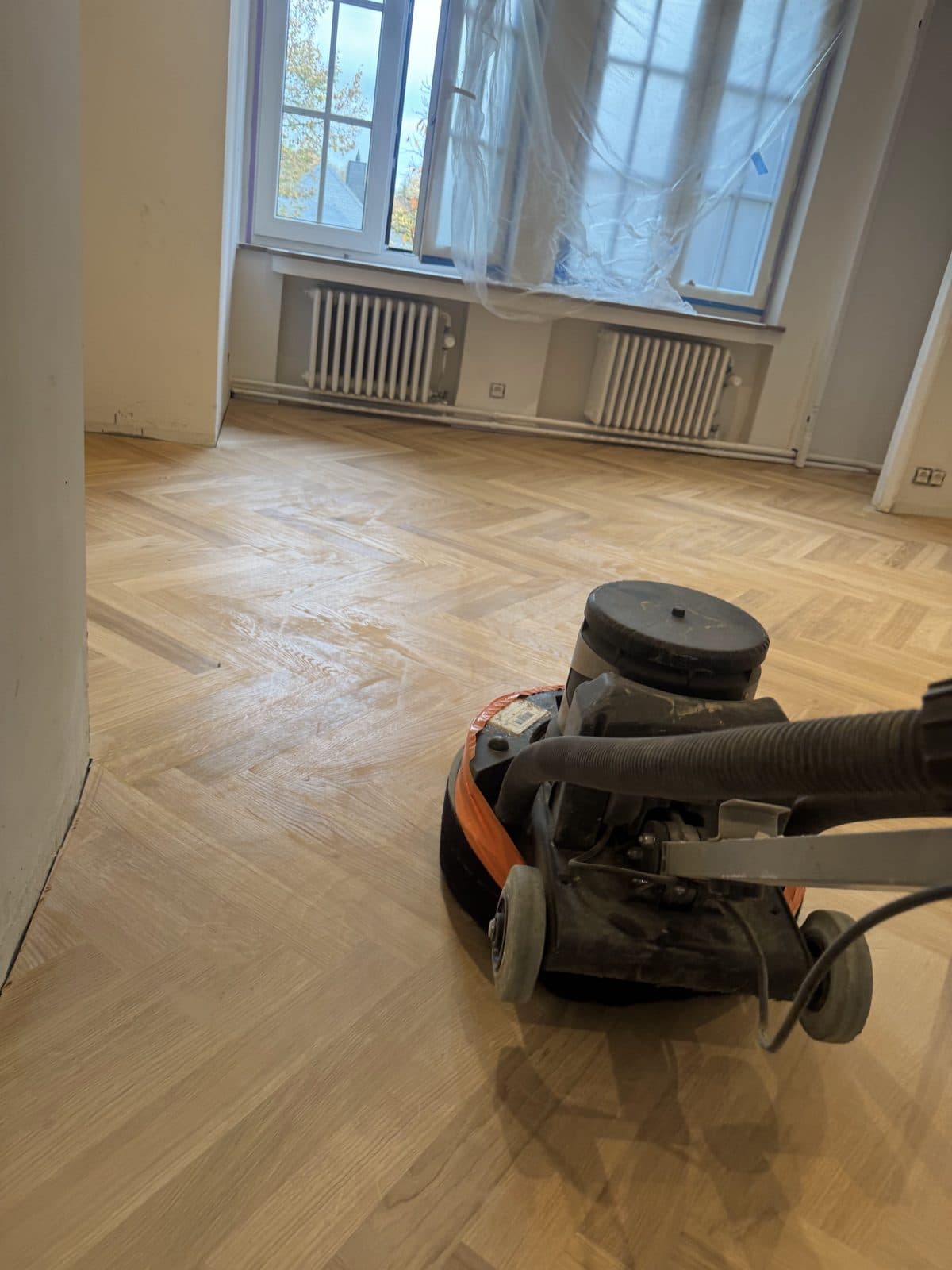 Ponceuse à parquet et son tuyau d’aspiration sur un sol à bâtons rompus poncé, fenêtre protégée par un film