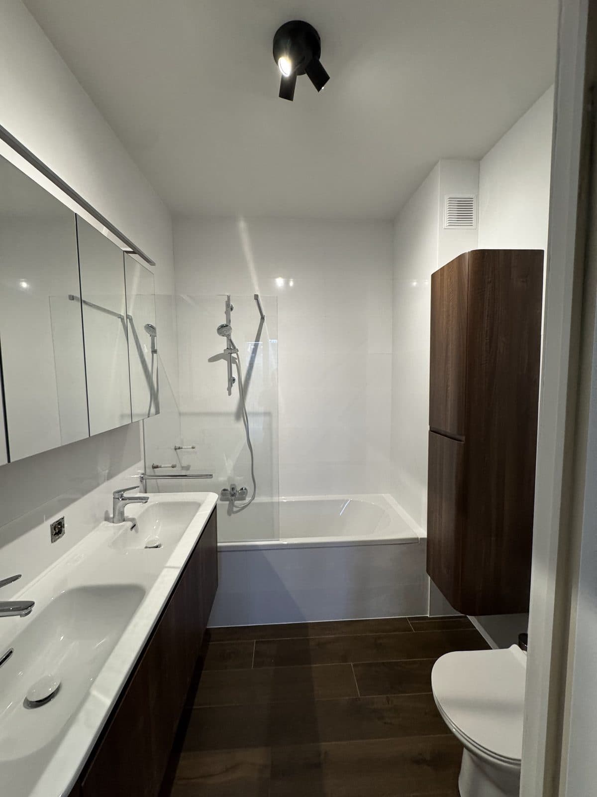 Salle de bain en carrelage blanc brillant : baignoire avec douche, colonne arrondie en bois sombre, double vasque, sol aspect bois foncé