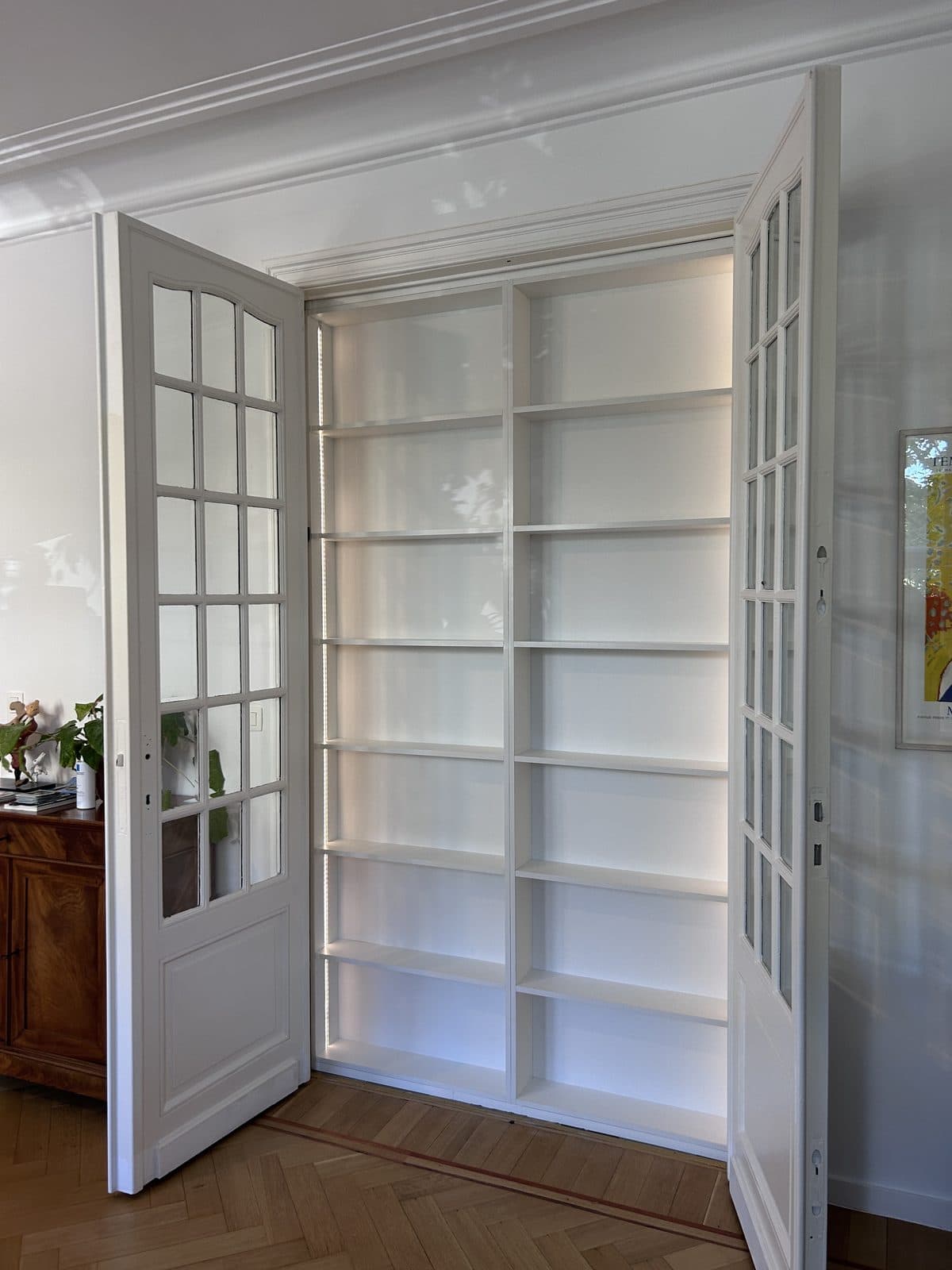 Bibliothèque blanche à éclairage latéral intégrée dans une embrasure, double porte vitrée ouverte, parquet à bâtons rompus