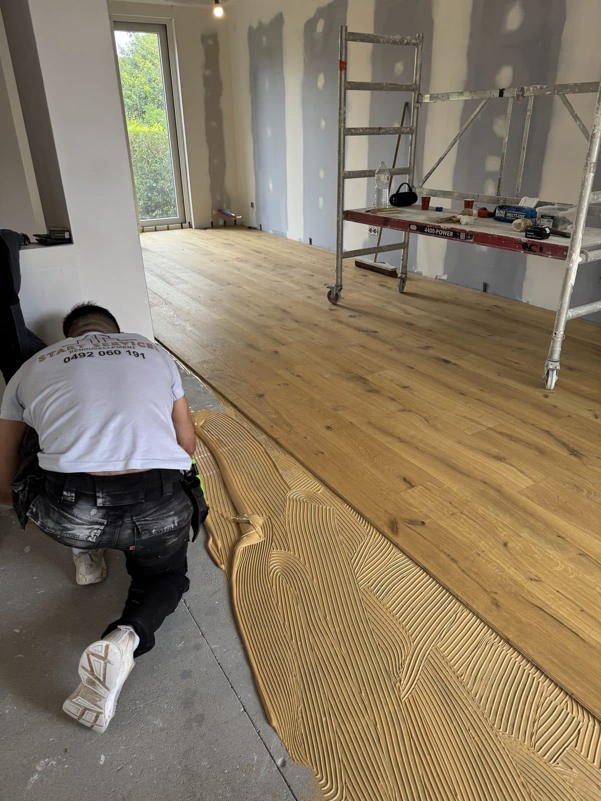 Un ouvrier à genoux, de dos, étale la colle à la spatule crantée devant le parquet en chêne à moitié posé