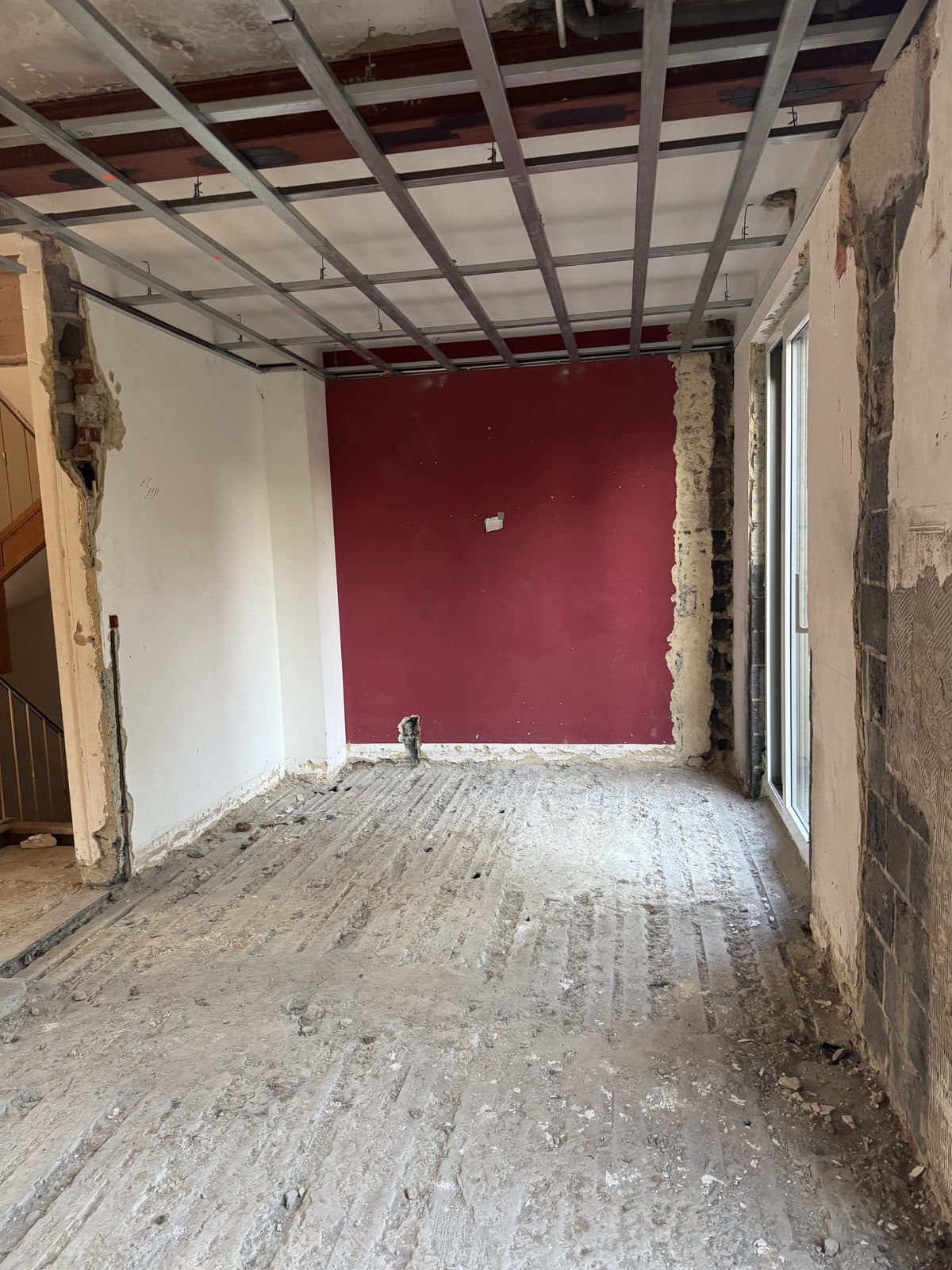 Pièce mise à nu : mur du fond bordeaux, ossature métallique au plafond, fenêtre coulissante à droite