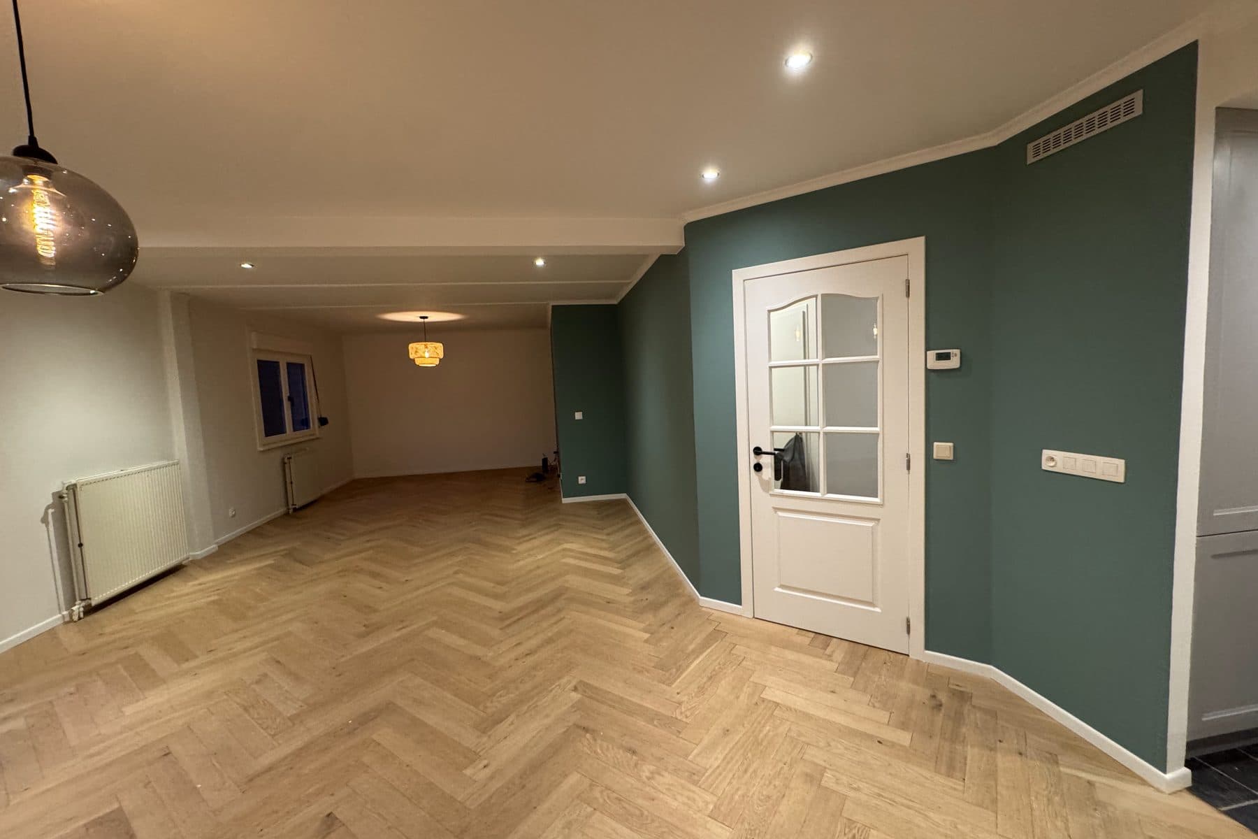 Longue pièce au mur vert profond, porte blanche à petits carreaux, parquet en chêne à bâtons rompus