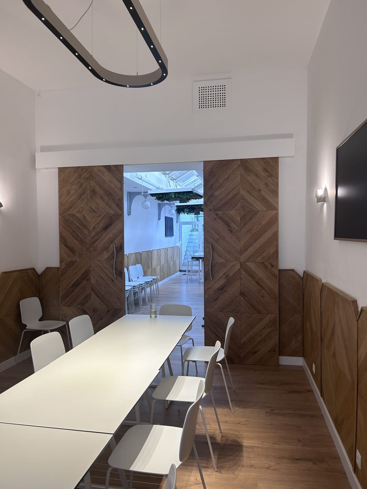 Salle de réunion : longue table blanche, portes en chêne à chevrons ouvertes sur un couloir vitré, soubassement en chêne